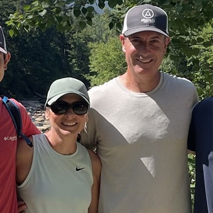 Man and woman outdoors wearing caps and sunglasses, smiling for a photo with a scenic forest background.