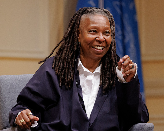 Whoopi Goldberg smiling during a public event, wearing a dark blazer and white shirt with long dreadlocks.