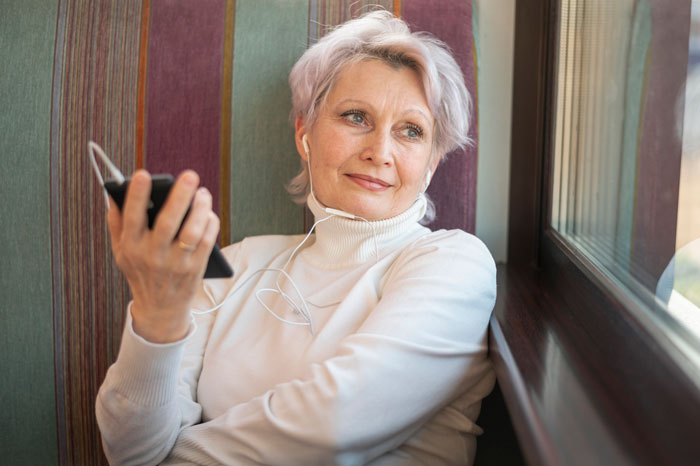 Older woman in turtleneck listening to music with earbuds, holding phone and staring thoughtfully out the window. Older woman in turtleneck listening to music with earbuds, holding phone and staring thoughtfully out the window.