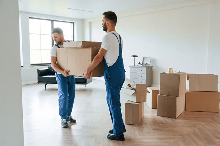 Two men in overalls carrying a large box into a mostly empty home, symbolizing husband&rsquo;s sudden decision to move in disabled siblings.