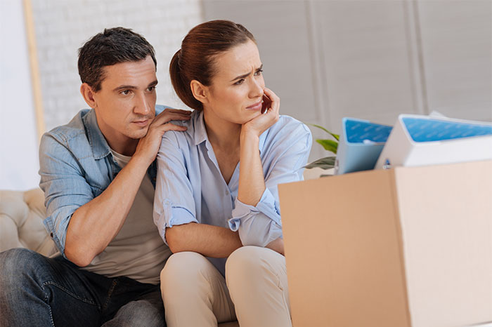 Worried wife and husband sit together in living room surrounded by moving boxes, facing marriage and family challenges.
