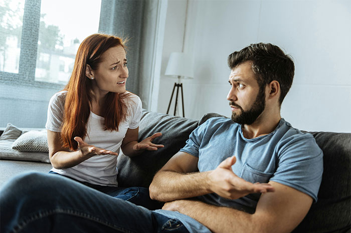 Wife and husband arguing on couch as husband's sudden decision to move in disabled siblings causes marital tension.