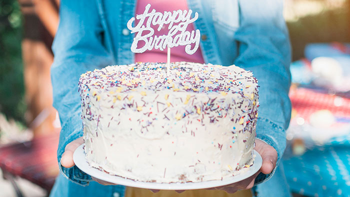 Person in denim jacket holding a birthday cake with sprinkles and a happy birthday topper outdoors in daylight - 7