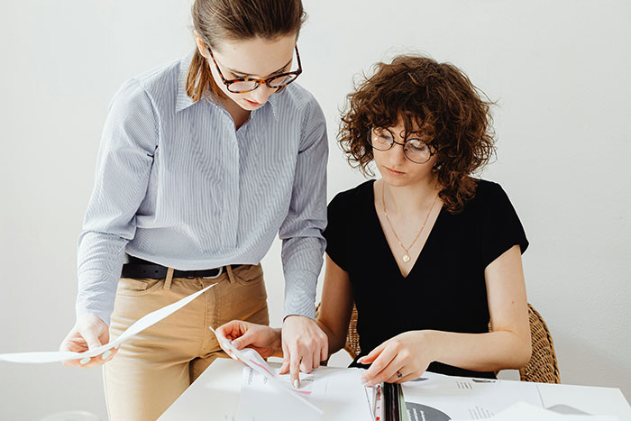 Two women reviewing documents at a desk, the annoyed worker showing frustration with a birthday cake nearby. - 13