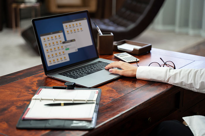 Worker at desk using laptop with documents and glasses nearby, appearing annoyed while avoiding birthday cake celebration. - 1