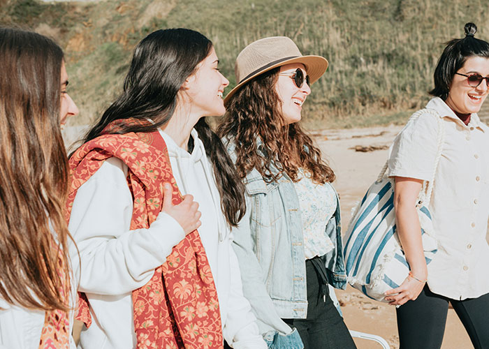 Four women walking and laughing outdoors on a sunny beach day, enjoying time together in casual outfits. - 13