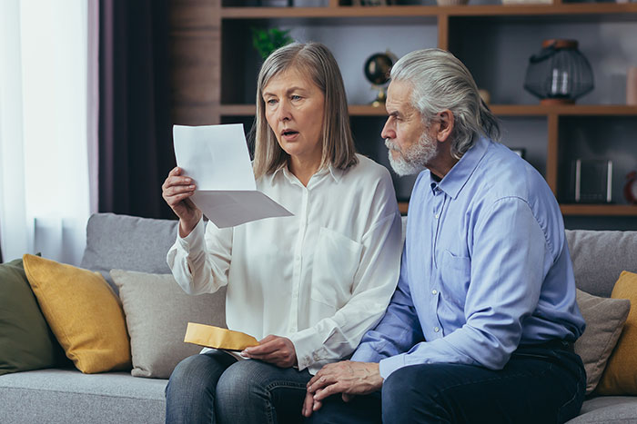 Middle-aged in-laws sitting on a couch, looking shocked while reading a letter about woman quitting her job. - 1