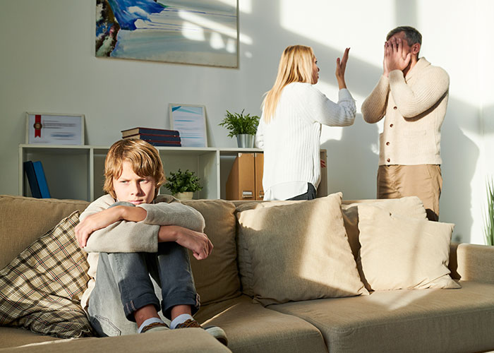 Co-parents arguing intensely in living room while upset child sits alone on couch, highlighting co-parenting misunderstanding issues.