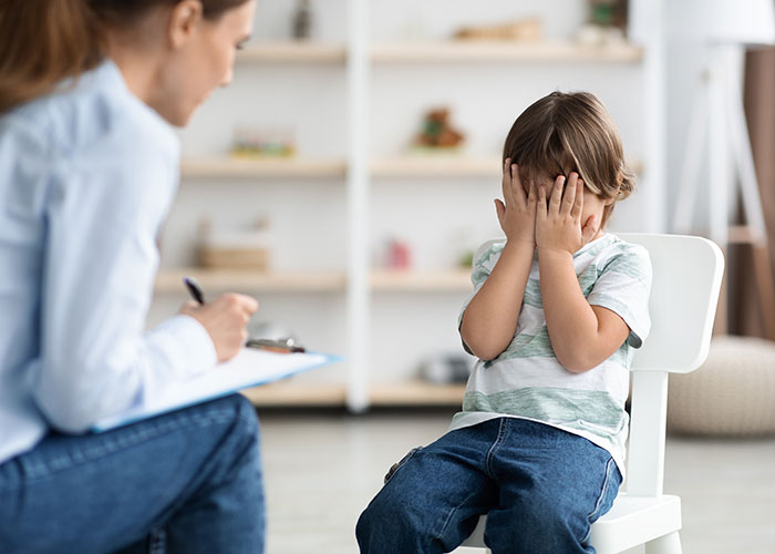 Young boy covering his face while a counselor takes notes during a co-parenting misunderstanding involving CPS intervention.