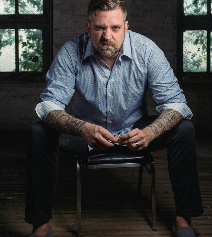 Man with tattoos sitting on a chair in a dim room, showing emotions related to cutting off a friend in abusive marriage.
