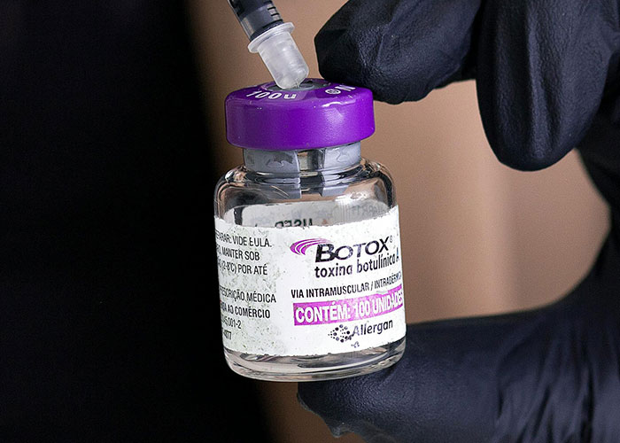 Gloved hand holding a Botox vial with needle, illustrating cosmetic procedures related to women getting facelifts at a young age. Gloved hand holding a Botox vial with needle, illustrating cosmetic procedures related to women getting facelifts at a young age.