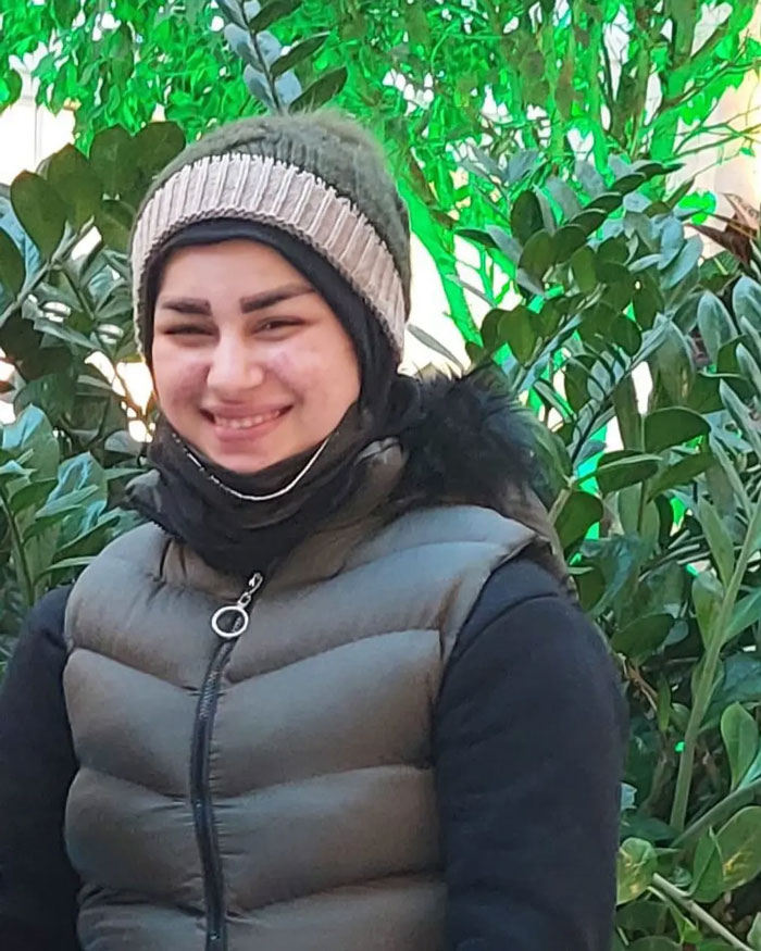 Smiling young woman wearing a winter hat and puffer vest standing outdoors with green leafy plants in the background.