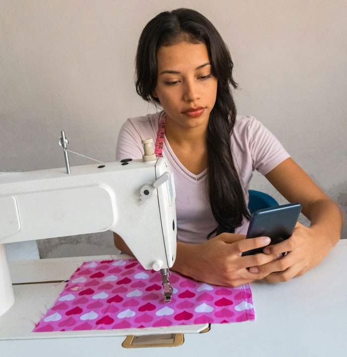 Young woman with sewing machine and fabric, looking at phone, symbolizing refusal to wear niece&rsquo;s handmade dress.