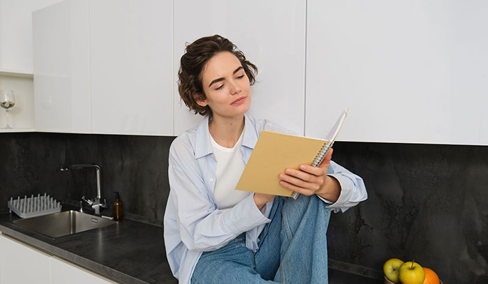 Young woman sitting in kitchen, looking thoughtfully at notebook, depicting emotions related to inheritance and gaslighting issues.