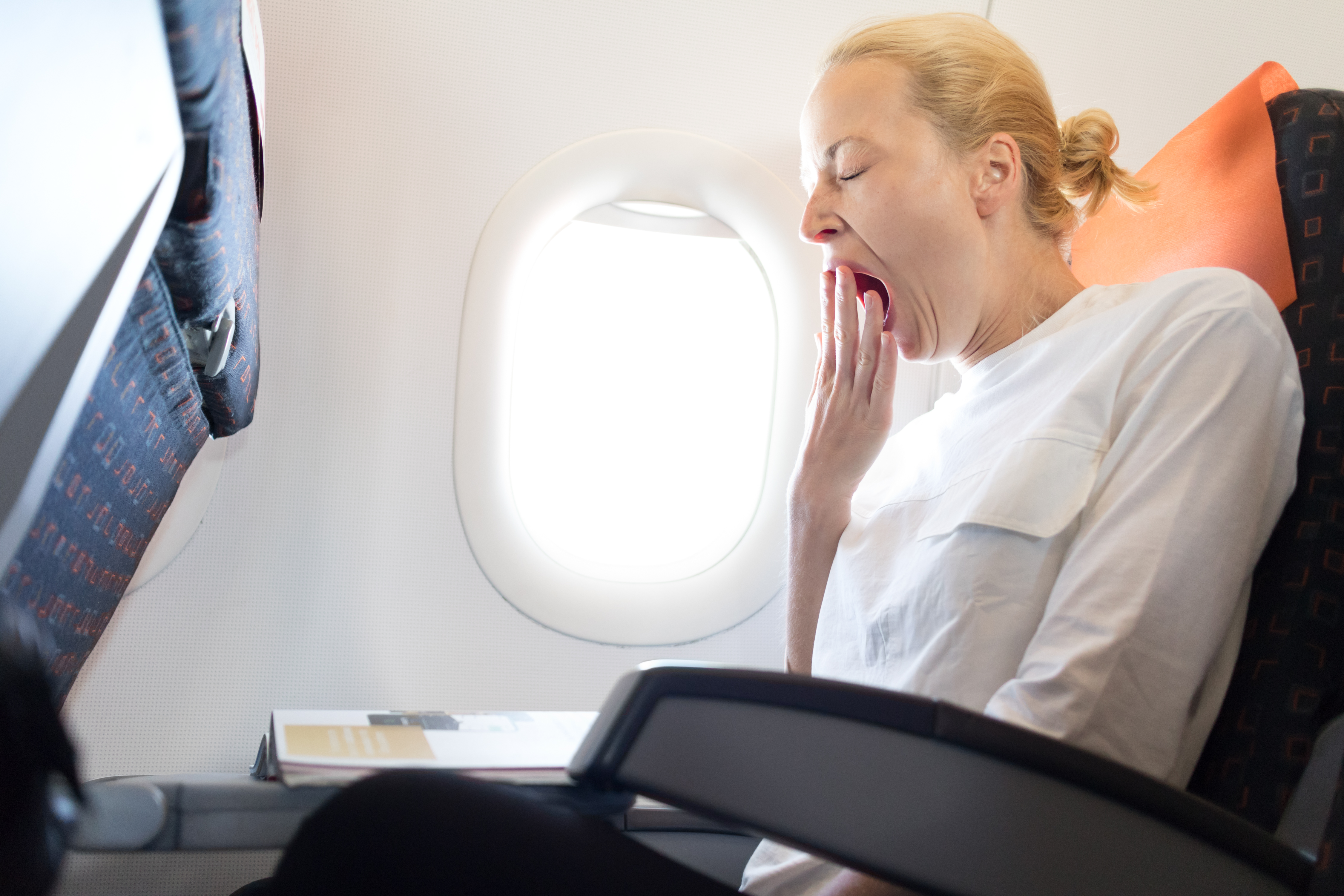 Woman yawning during flight, sitting by window while rowdy kids and drunk parents are nearby on plane. - 1