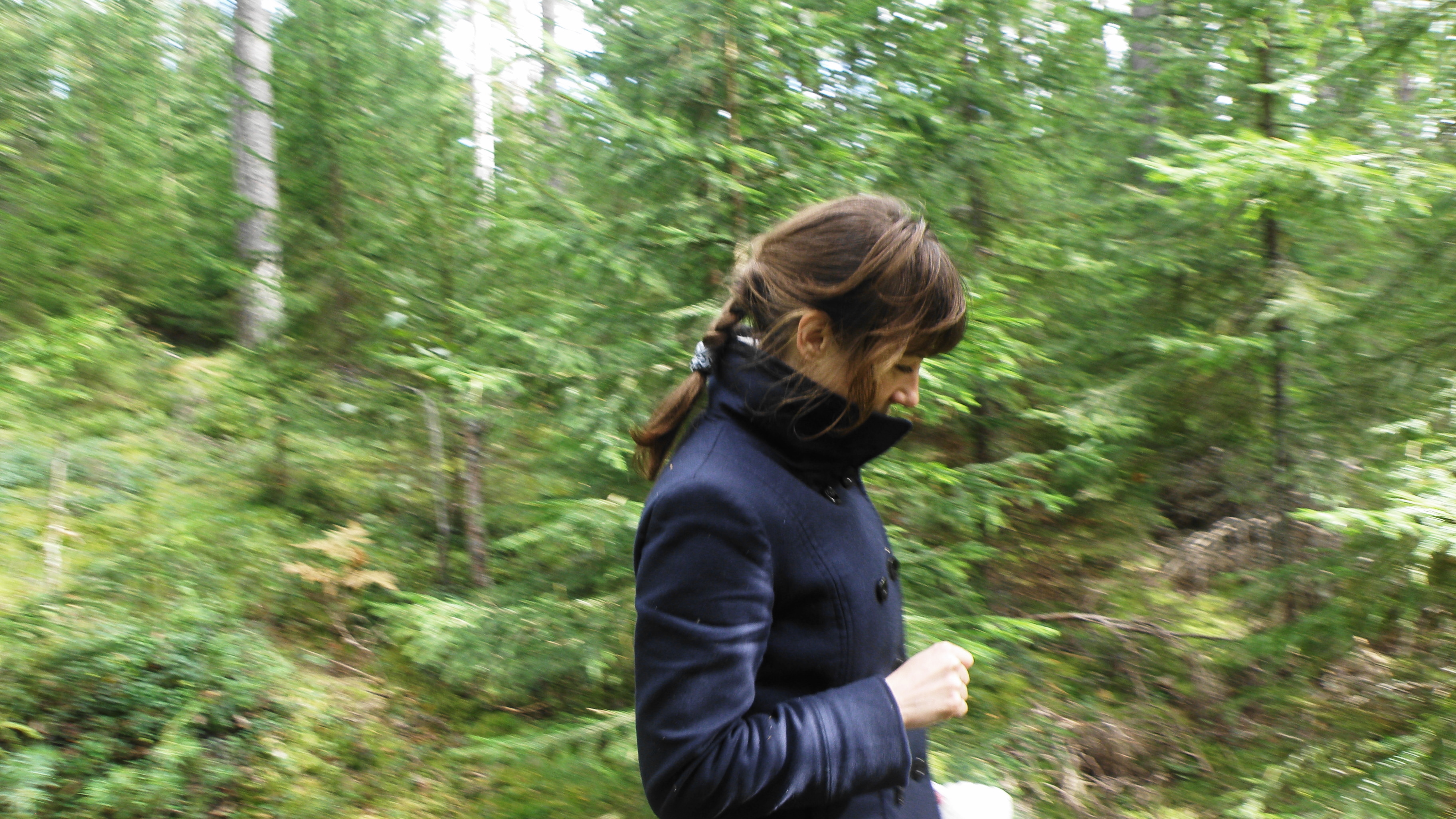 Woman with braided hair wearing a dark coat walking alone in dense green woods, appearing followed by strange man nearby.