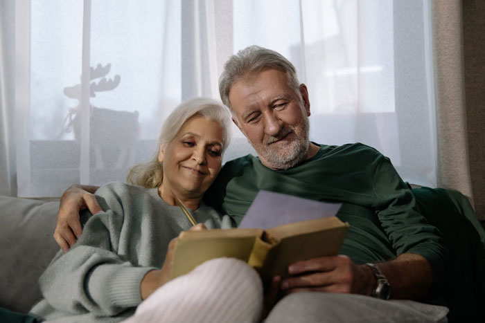 Older woman and man sitting on a couch together, smiling and reading a book, depicting a couple facing a difficult decision.