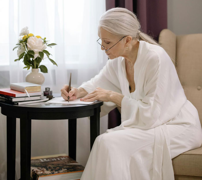 Older woman in white robe writing a letter at a table, reflecting on leaving her partner homeless after she dies