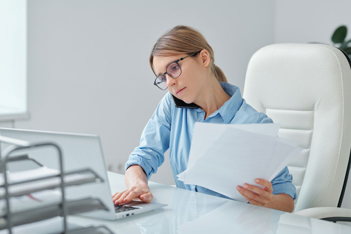 Female supervisor multitasking with phone, papers, and laptop, looking concerned while working in a modern office setting.