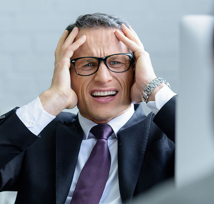 Frustrated businessman in glasses holding head, reacting to penalty for being late in a corporate office setting.