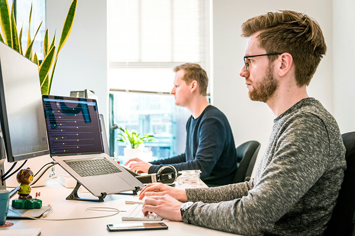 Two men working at their desks on computers in a bright office, highlighting penalty for being late at work.