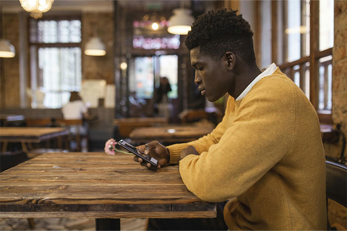Young man in a cafe, wearing a mustard sweater, looking at his phone while servers share the worst dates at work.