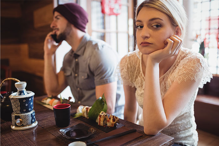 Young woman looking bored at a restaurant table while her date is distracted, illustrating servers sharing worst dates witnessed at work.