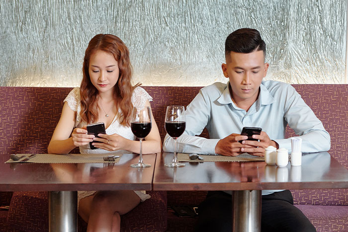 A couple on a date at a restaurant, both distracted by their phones, illustrating servers sharing worst dates witnessed at work.