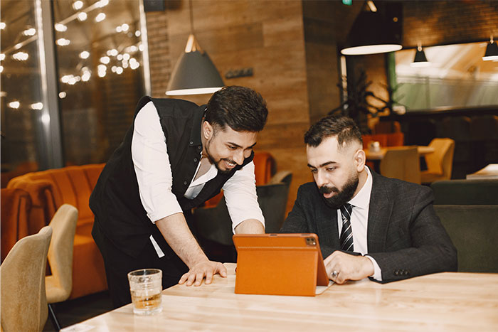 Server in a vest showing a man in a suit something on a tablet at a restaurant while sharing worst dates witnessed at work.