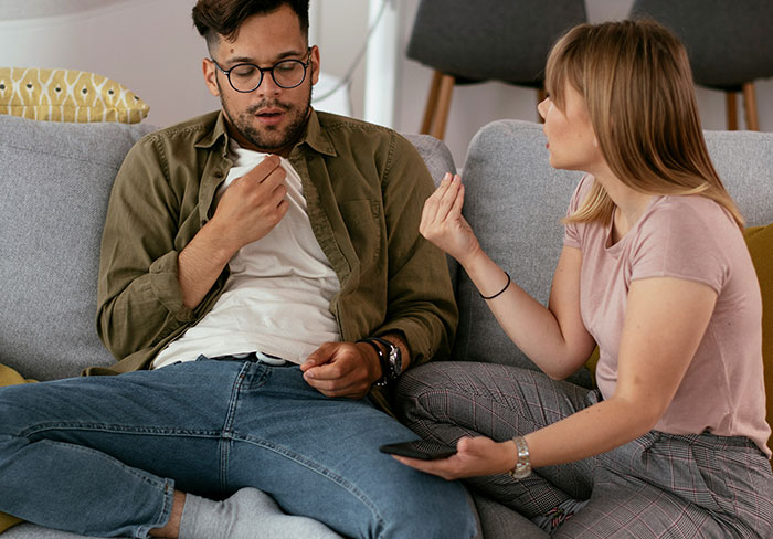 Man trying to mansplain something to a woman who looks unimpressed and is rolling her eyes on a couch.