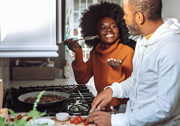 Couple cooking together in kitchen, woman reacting with a smile while man mansplains something nearby.