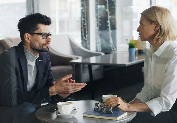 Man trying to mansplain to woman at a cafe table while she listens and rolls her eyes during the conversation.