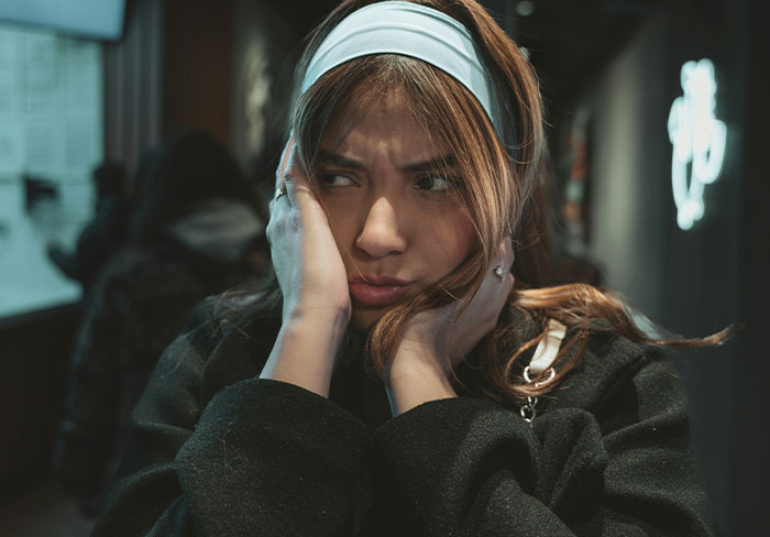 Young woman with a frustrated expression covering her ears, reacting to mansplain attempts in a dimly lit setting.