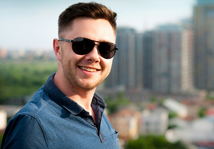 Young man wearing sunglasses and a blue shirt smiling outdoors, illustrating mansplain scenarios with women rolling their eyes.