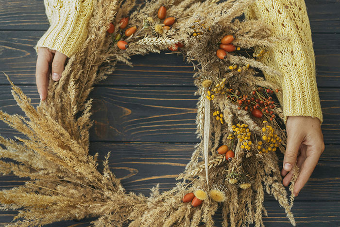 Hands in a yellow sweater arranging a decorative dried flower wreath on a dark wooden surface.