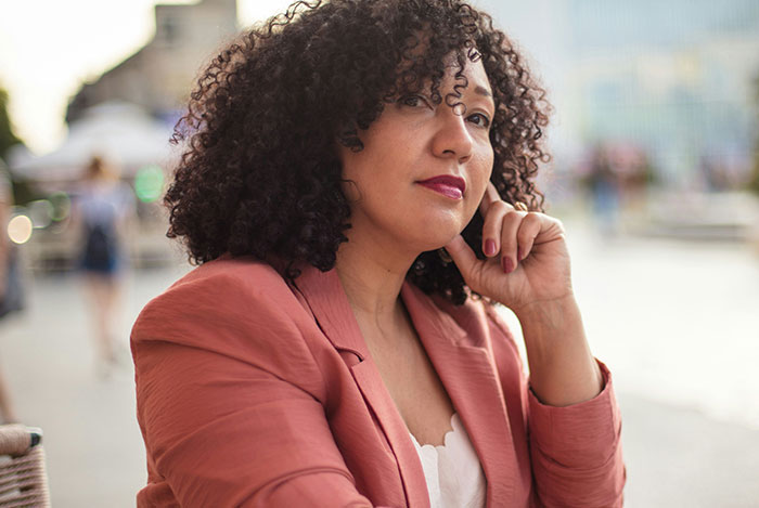 Woman with curly hair wearing a coral blazer, showing an expression of eye-rolling in response to mansplaining outdoors.