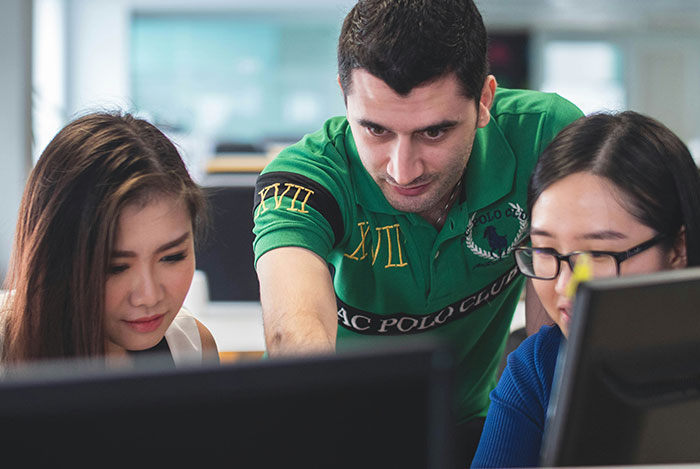 Man explaining something to two women at computers while they listen and roll their eyes in an office setting.