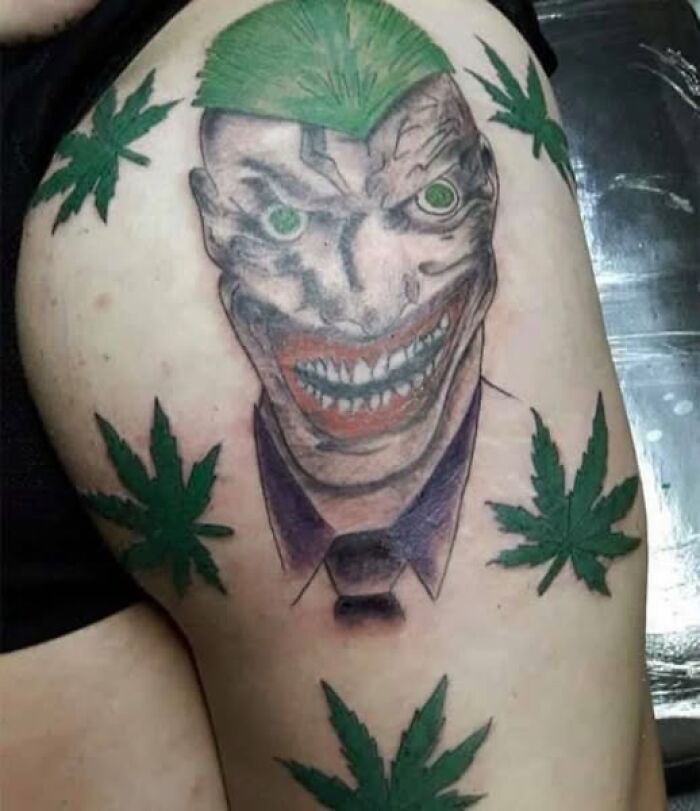 Tattoo of a distorted Joker face with green hair surrounded by poorly drawn green leaves on upper arm, example of bad tattoos.