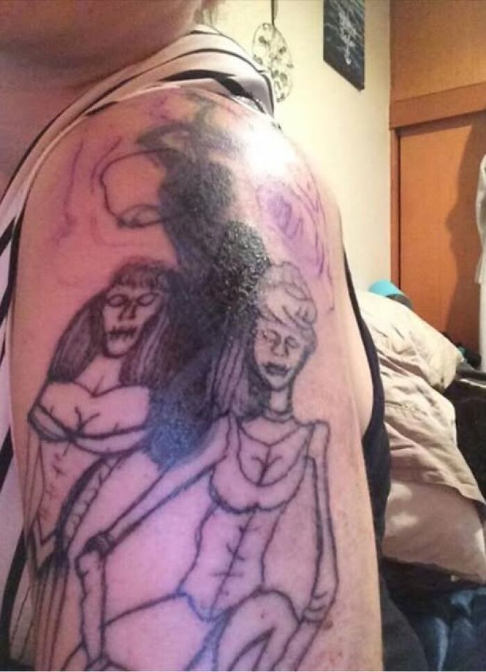 Abysmally bad tattoo of two women with distorted features on a person’s upper arm in a casual indoor setting.