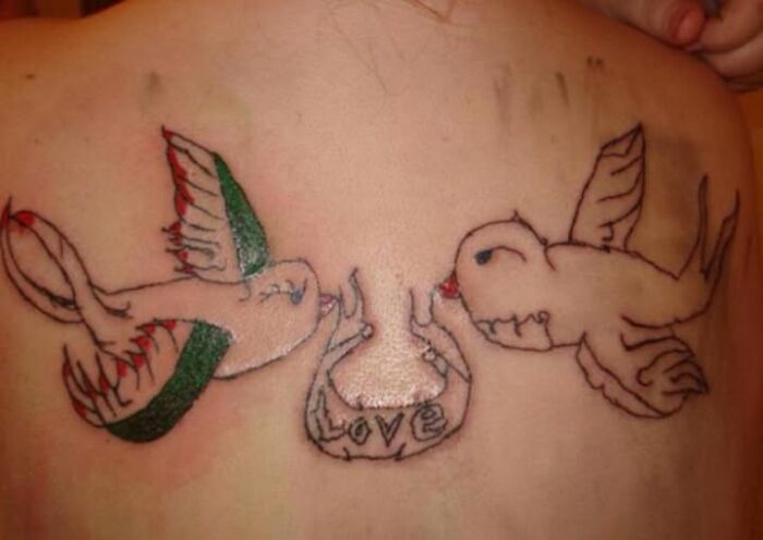 Abysmally bad tattoo of two birds with a heart and the word love, poorly drawn and inked on a person's back.
