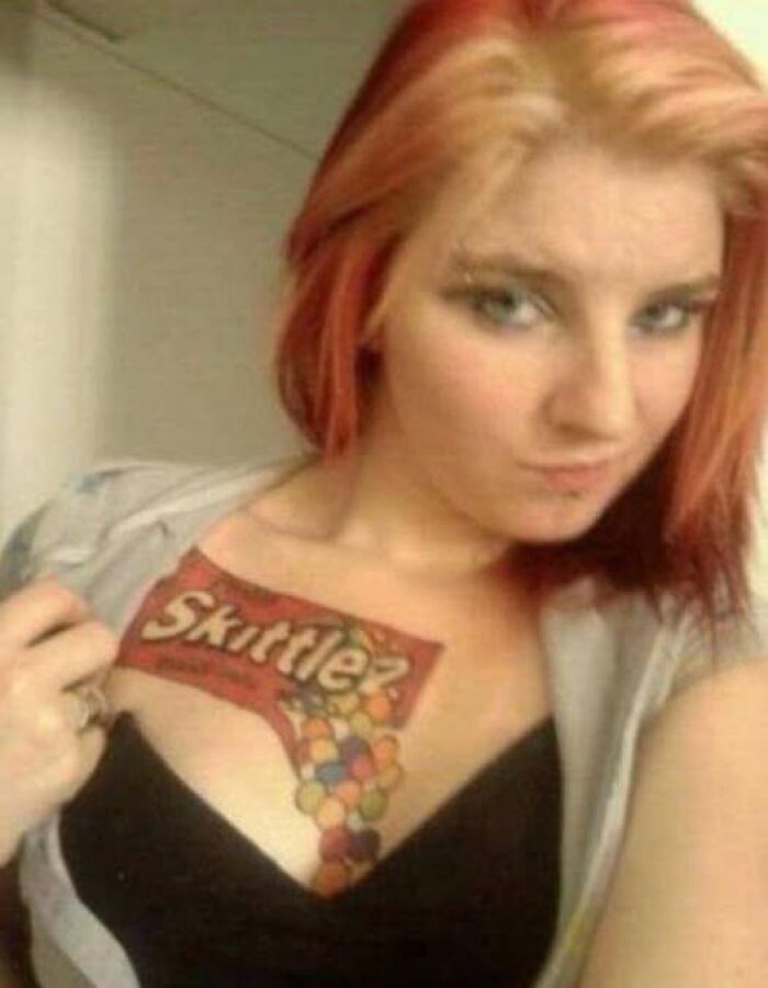 Young woman showing a large Skittles tattoo on her chest, an example of abysmally bad tattoos featured on dedicated FB pages.