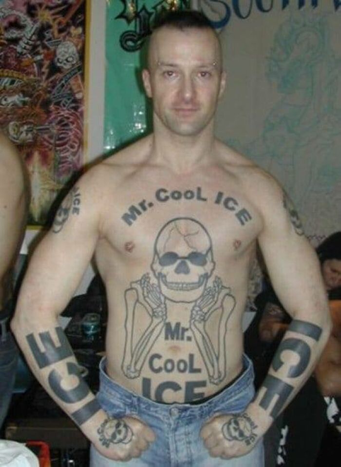 Man with abysmally bad tattoos featuring large Mr. Cool Ice skull designs on chest, arms, and knuckles at a social gathering.