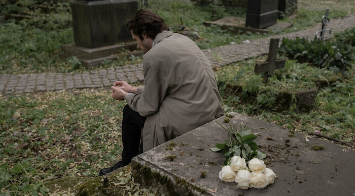 Man in a trench coat sitting alone on a gravestone in a cemetery, reflecting quietly in a somber mood.