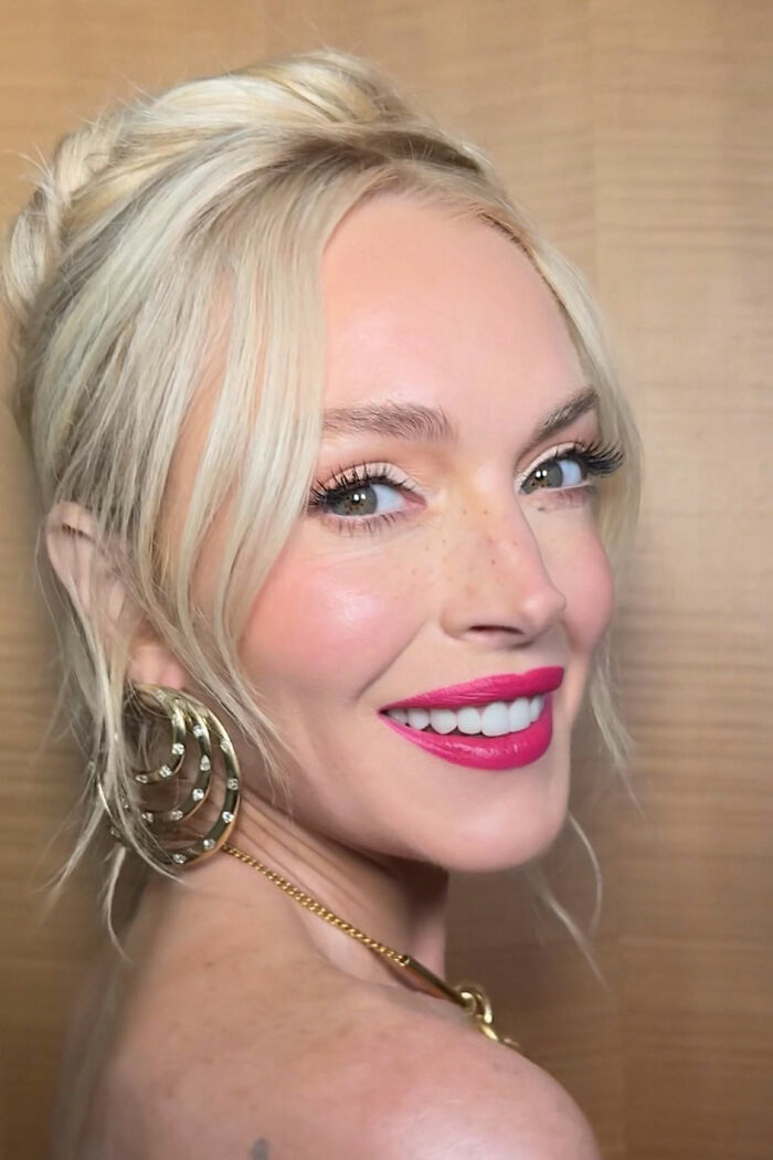 Lindsay Lohan smiling with fresh-faced glow, blonde hair, bold pink lipstick, and gold hoop earrings in close-up portrait.