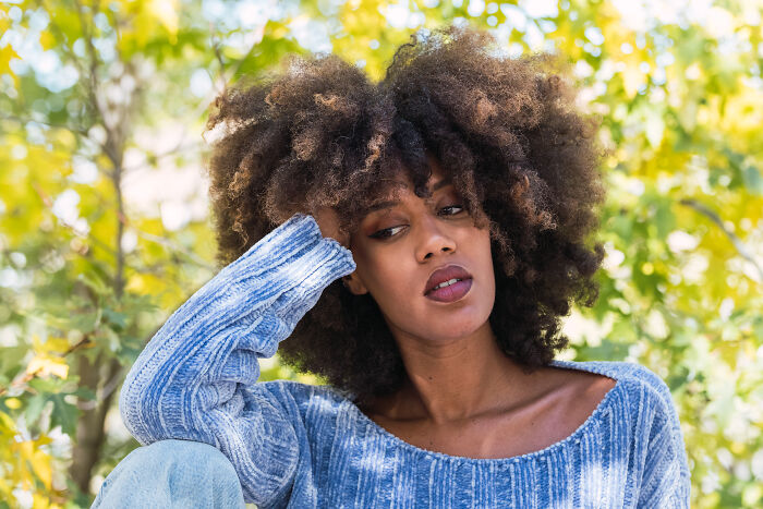 Woman with curly hair wearing a blue sweater, outdoors surrounded by green leaves, reflecting on normal people’s habits. - 25
