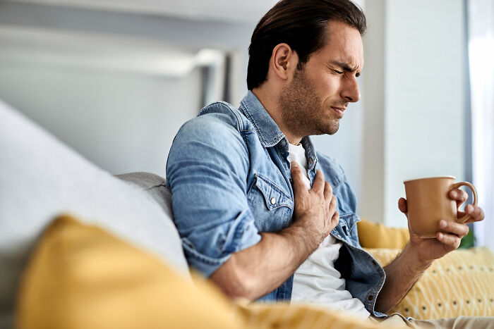 Man in a denim shirt sitting on couch holding mug and clutching chest, depicting impostor patients needing less meds and common sense