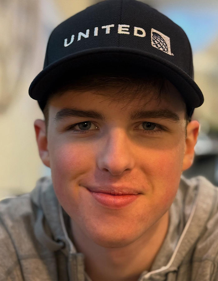 Teenage Young Sheldon child star wearing a black United cap and gray hoodie, smiling in a close-up portrait.