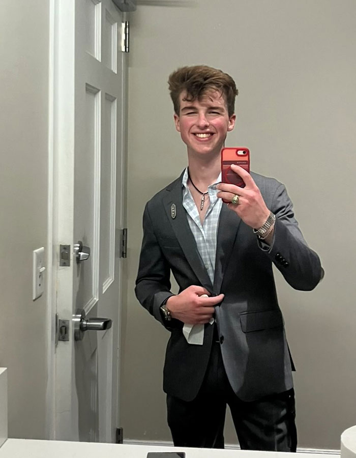 Young Sheldon child star at 17 wearing a suit, smiling and taking a mirror selfie in a bathroom.