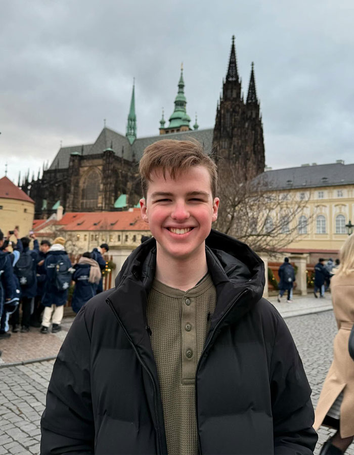 Young Sheldon child star at 17, smiling outdoors in a black jacket with historic buildings in the background.