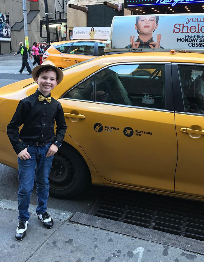 Young Sheldon child star in black shirt and bow tie standing next to a yellow taxi in an urban setting.